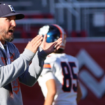 Man wearing a ball cap on a football field talking to players